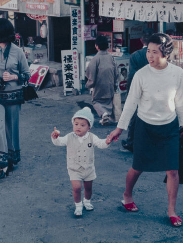 A woman holds a young child's hand while walking through a busy street market, captured in a vintage colour photograph