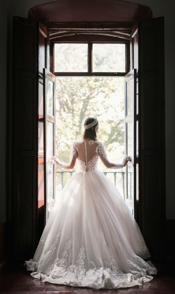 Bride in an elegant lace wedding gown standing by an open window, backlit by soft natural light in a romantic bridal setting.