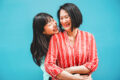 An Asian mother and daughter laughing and embracing outdoors against a teal background