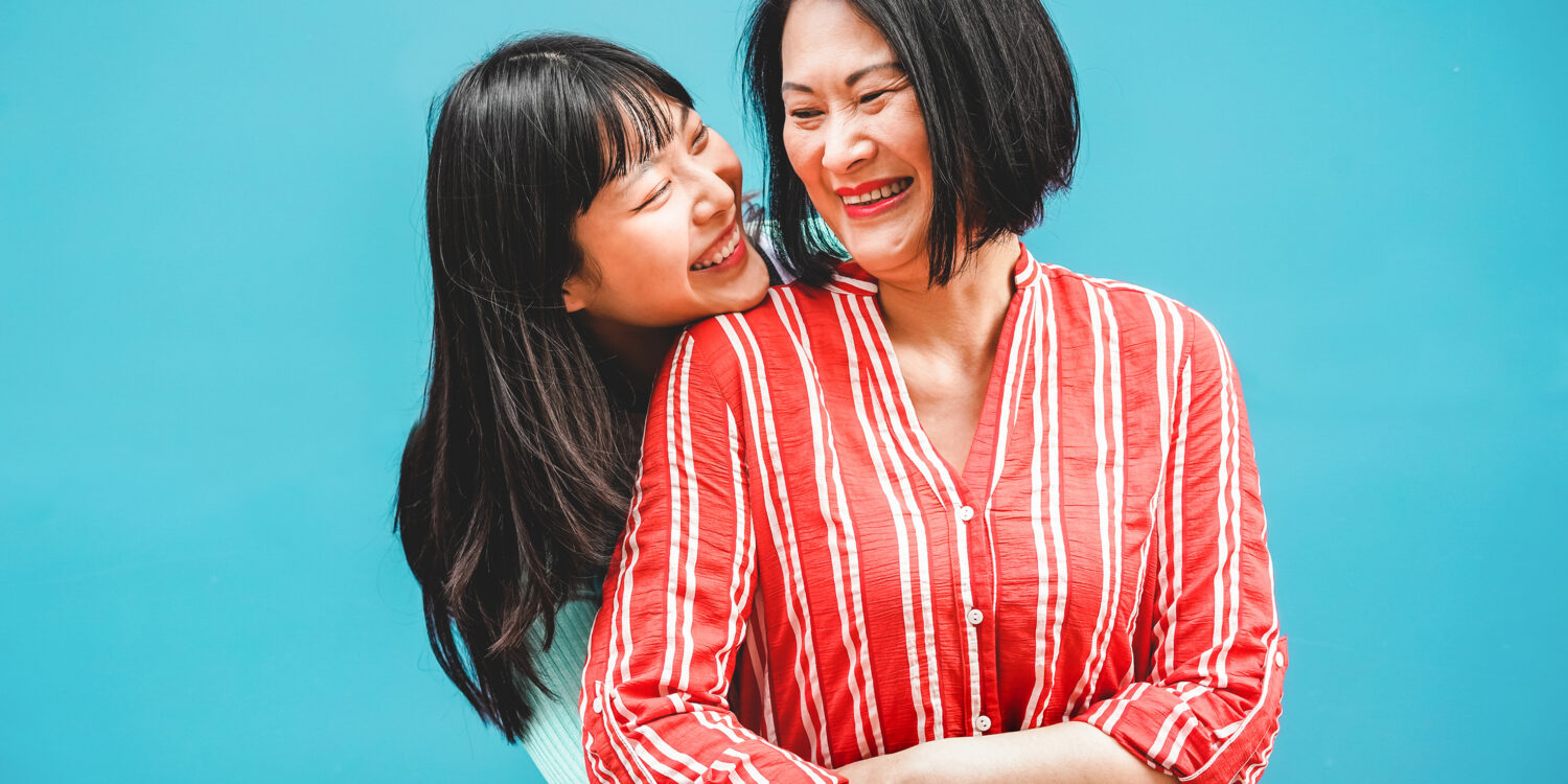 An Asian mother and daughter laughing and embracing outdoors against a teal background