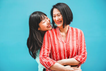 An Asian mother and daughter laughing and embracing outdoors against a teal background