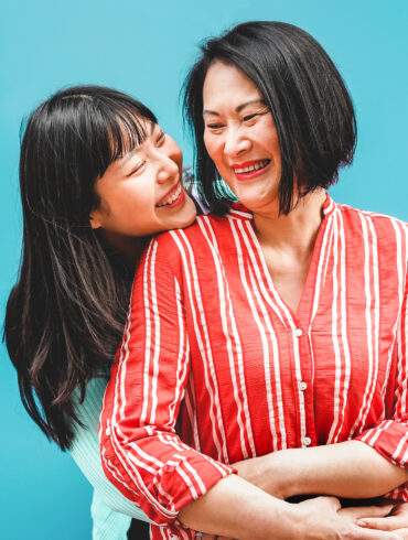 An Asian mother and daughter laughing and embracing outdoors against a teal background