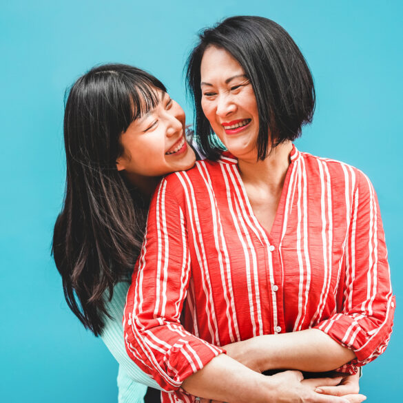 An Asian mother and daughter laughing and embracing outdoors against a teal background