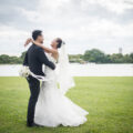 Happy bride and groom embracing on a grassy field by a lake during an outdoor wedding photoshoot.