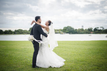 Happy bride and groom embracing on a grassy field by a lake during an outdoor wedding photoshoot.