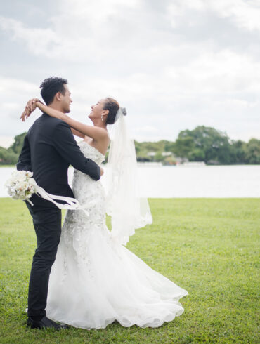 Happy bride and groom embracing on a grassy field by a lake during an outdoor wedding photoshoot.