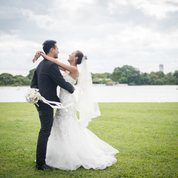 Happy bride and groom embracing on a grassy field by a lake during an outdoor wedding photoshoot.