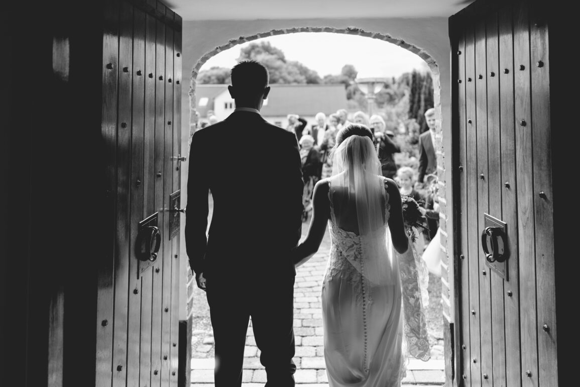 Bride and groom entering a wedding ceremony through large wooden doors, with guests gathered outside in a classic black and white scene.