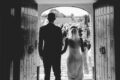 Bride and groom entering a wedding ceremony through large wooden doors, with guests gathered outside in a classic black and white scene.