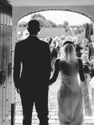 Bride and groom entering a wedding ceremony through large wooden doors, with guests gathered outside in a classic black and white scene.