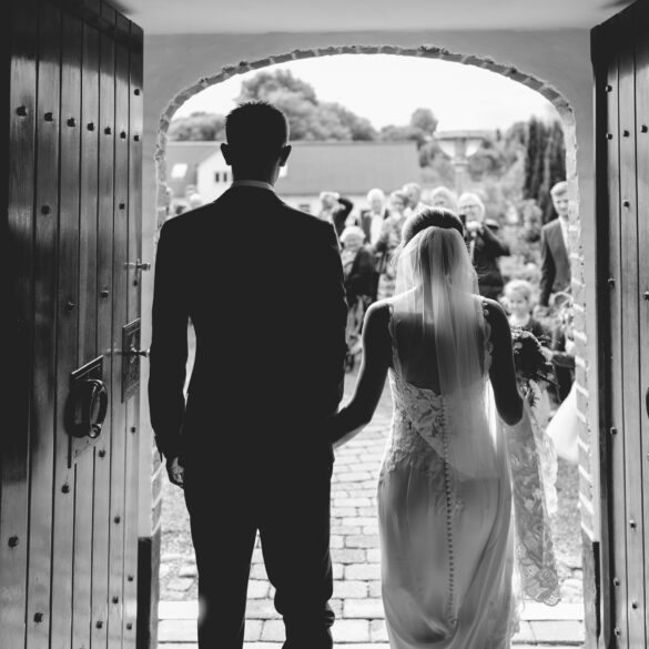 Bride and groom entering a wedding ceremony through large wooden doors, with guests gathered outside in a classic black and white scene.