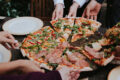 A group of people reaching for slices of pizza topped with prosciutto, salami, arugula and roasted peppers on a wooden table, representing the new restaurants and dining openings in Singapore this April 2026.