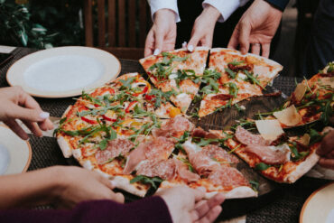 A group of people reaching for slices of pizza topped with prosciutto, salami, arugula and roasted peppers on a wooden table, representing the new restaurants and dining openings in Singapore this April 2026.