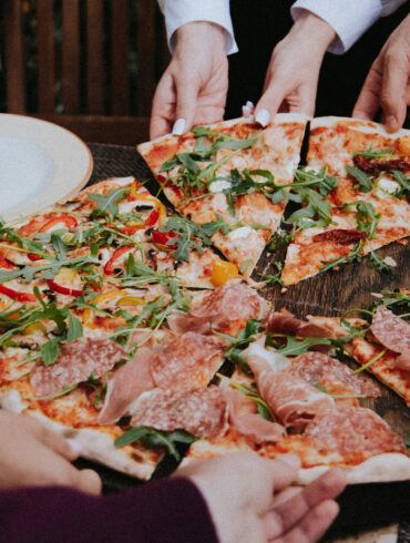 A group of people reaching for slices of pizza topped with prosciutto, salami, arugula and roasted peppers on a wooden table, representing the new restaurants and dining openings in Singapore this April 2026.