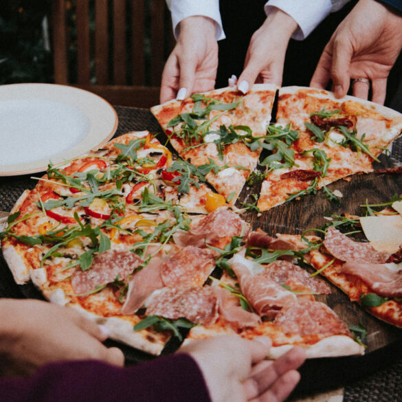 A group of people reaching for slices of pizza topped with prosciutto, salami, arugula and roasted peppers on a wooden table, representing the new restaurants and dining openings in Singapore this April 2026.