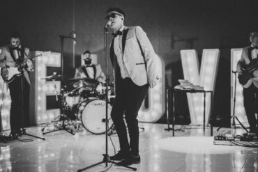 Wedding live band performing on stage with singer and musicians in formal attire, entertaining guests at a reception with illuminated ‘LOVE’ letters.