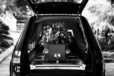 Black-and-white image of a hearse with its rear door open, revealing a wooden coffin topped with a floral arrangement inside.