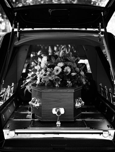Black-and-white image of a hearse with its rear door open, revealing a wooden coffin topped with a floral arrangement inside.