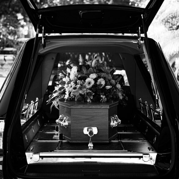 Black-and-white image of a hearse with its rear door open, revealing a wooden coffin topped with a floral arrangement inside.
