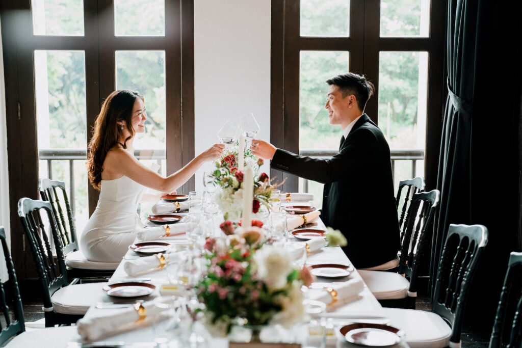A couple toasting at a elegantly set long dining table inside The Alkaff Mansion, with lush greenery visible through the windows