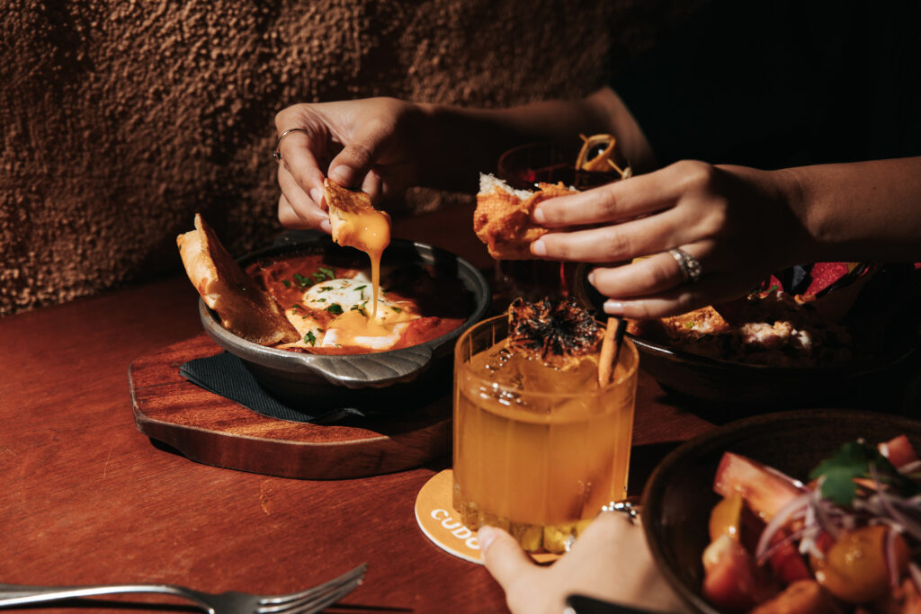 Hands tearing into a skillet of Rancheros baked eggs at CUDO, with crusty bread for dipping into the spiced tomato stew and a cocktail garnished with charred citrus on the side.