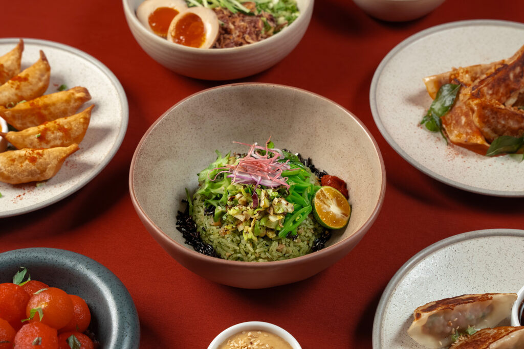 A spread of dishes at Dumpling Darlings New Bahru, including pan-fried and crispy dumplings, a green rice bowl topped with charred vegetables, marinated egg with pulled meat, and pickled cherry tomatoes.