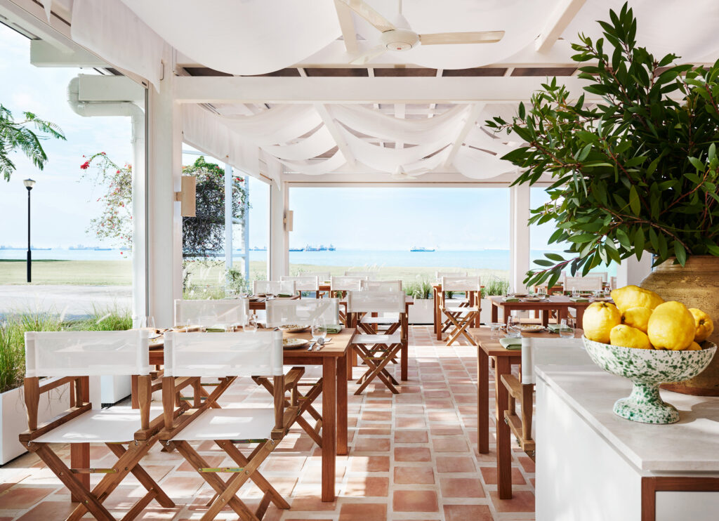 The sun-drenched alfresco terrace at Fico, East Coast Park, with terracotta tiles, timber furniture, and sea views