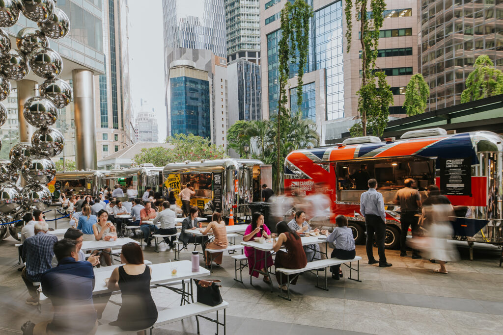Office workers gathered at communal benches in the covered plaza at Gourmet Park OFC, with the Union Jack-wrapped Carnaby Burger Bar truck and Jamaica Jerk Chicken Co. truck lining the back against the CBD skyline.