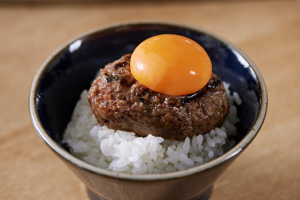 A signature hamburg steak from Hikiniku To Come, served on a bed of white rice in a dark ceramic bowl and topped with a glossy egg yolk.