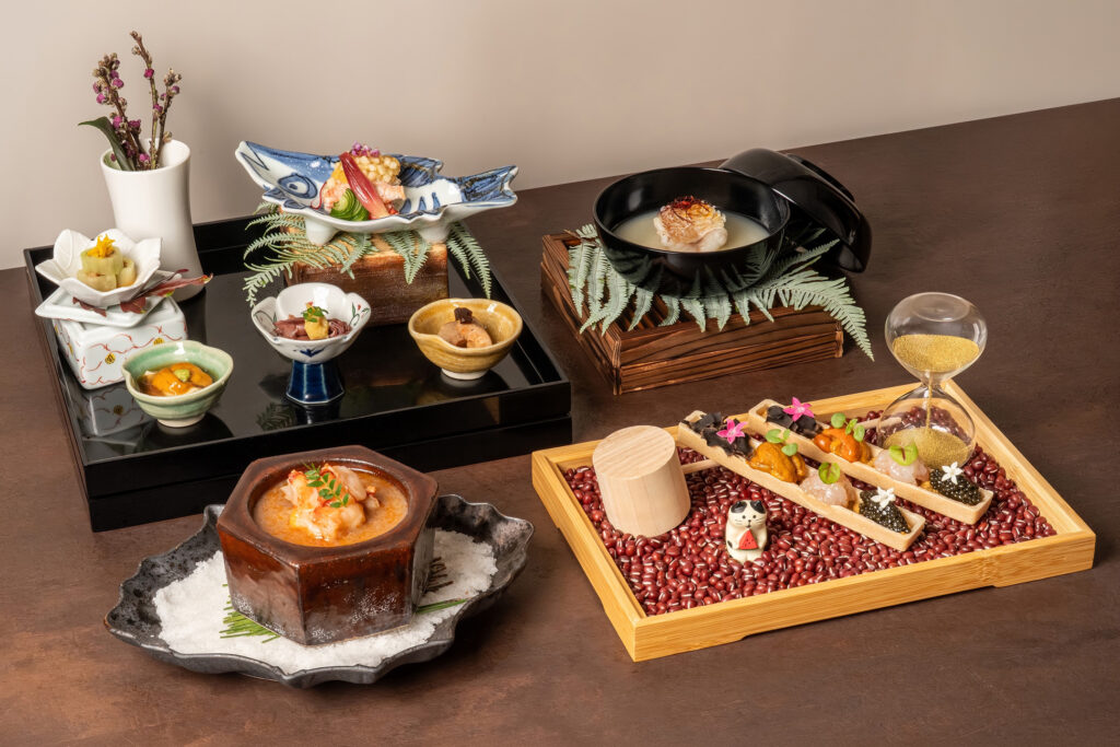 A spread of dishes at Jiin Omakase, including the seasonal appetiser on a fish-shaped plate, a chawanmushi-style soup in a black lidded bowl, a hot pot in a hexagonal vessel, and the signature Monaka course presented on a bed of adzuki beans.