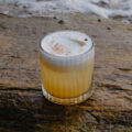 A foamy sour cocktail with a dusting of spice, served in a ribbed glass on a wet coastal rock with waves in the background.