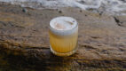 A foamy sour cocktail with a dusting of spice, served in a ribbed glass on a wet coastal rock with waves in the background.