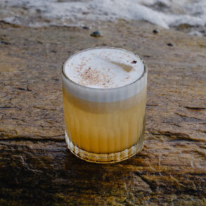 A foamy sour cocktail with a dusting of spice, served in a ribbed glass on a wet coastal rock with waves in the background.