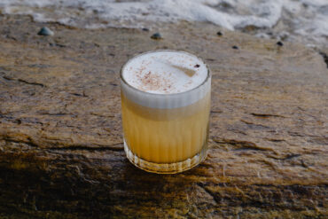 A foamy sour cocktail with a dusting of spice, served in a ribbed glass on a wet coastal rock with waves in the background.