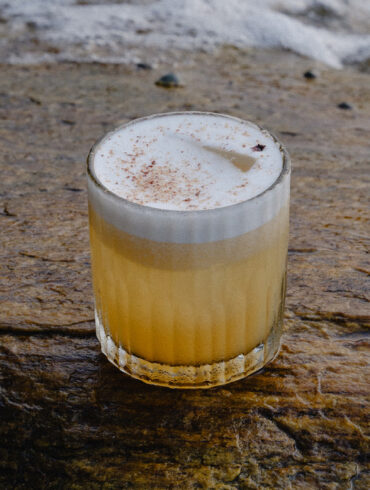 A foamy sour cocktail with a dusting of spice, served in a ribbed glass on a wet coastal rock with waves in the background.
