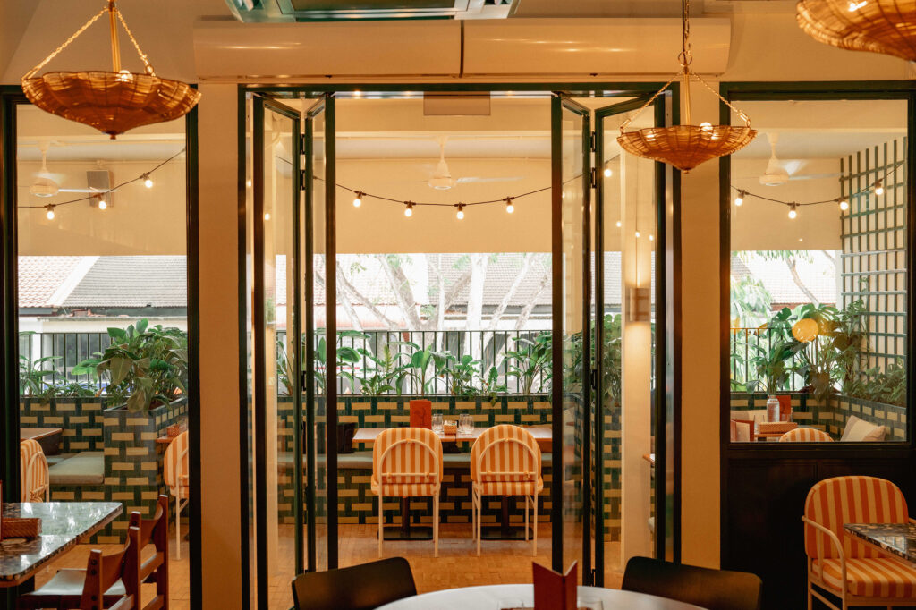 The interior of Merci Marcel Serangoon Gardens, with rattan pendant lights, string lighting, orange-and-cream striped chairs, and bifold windows opening onto a tiled terrace lined with plants.