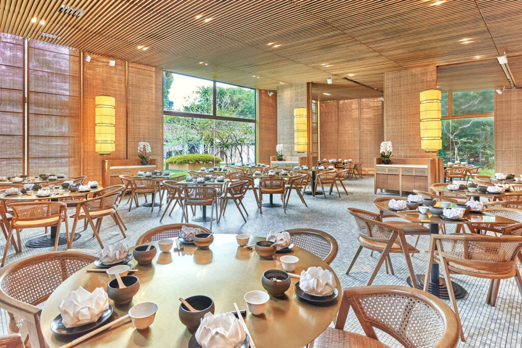The warm timber-lined interior of the main dining hall of Min Jiang at Dempsey, surrounded by greenery through floor-to-ceiling windows