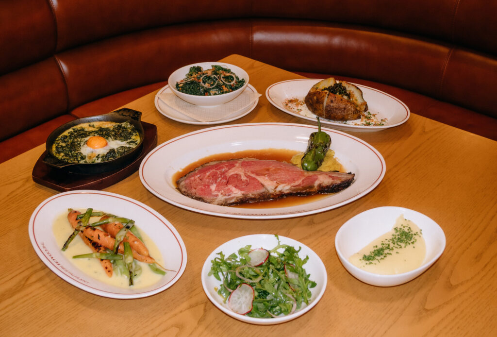 The Sunday Prime Rib spread at The Coach Restaurant, with a sliced cut of prime rib at the centre surrounded by sides including creamed spinach with egg yolk, kale salad, a baked russet potato topped with caviar, glazed carrots, arugula salad, and a side of sauce.