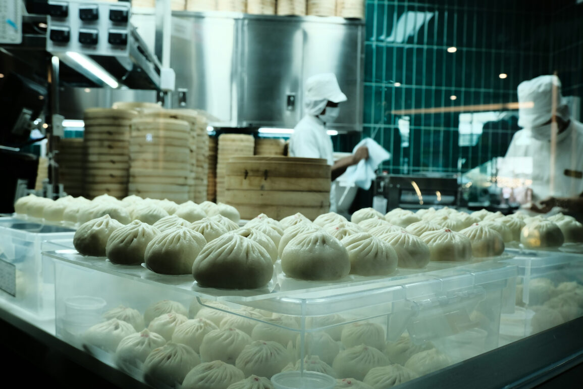 Trays of freshly steamed dumplings on display in a busy kitchen, with chefs working behind a glass partition stacked with bamboo steamers.