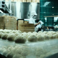 Trays of freshly steamed dumplings on display in a busy kitchen, with chefs working behind a glass partition stacked with bamboo steamers.