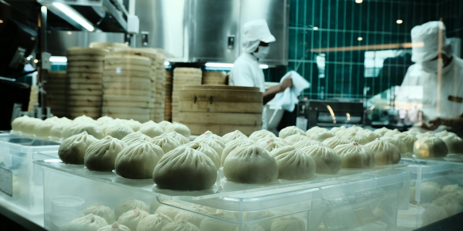 Trays of freshly steamed dumplings on display in a busy kitchen, with chefs working behind a glass partition stacked with bamboo steamers.