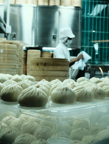 Trays of freshly steamed dumplings on display in a busy kitchen, with chefs working behind a glass partition stacked with bamboo steamers.