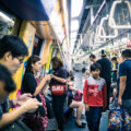 Commuters standing and seated inside a busy Singapore MRT carriage during peak hours, with overhead handrails and yellow interior panels visible.