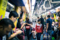 Commuters standing and seated inside a busy Singapore MRT carriage during peak hours, with overhead handrails and yellow interior panels visible.