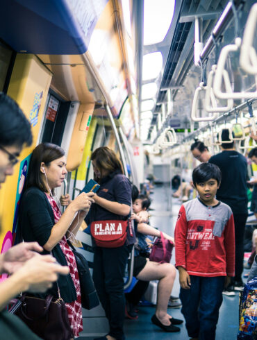 Commuters standing and seated inside a busy Singapore MRT carriage during peak hours, with overhead handrails and yellow interior panels visible.