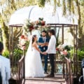 A couple exchanging vows at an outdoor garden wedding ceremony beneath a flower-adorned gazebo