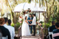 A couple exchanging vows at an outdoor garden wedding ceremony beneath a flower-adorned gazebo