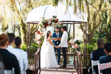 A couple exchanging vows at an outdoor garden wedding ceremony beneath a flower-adorned gazebo