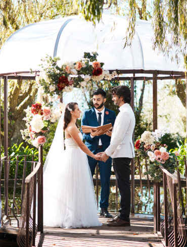 A couple exchanging vows at an outdoor garden wedding ceremony beneath a flower-adorned gazebo