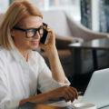 Woman in white shirt and glasses taking a phone call while working on a laptop at a cafe table with a coffee cup beside her.